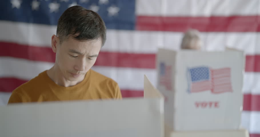 Frontal close up on Asian man casting votes in election. Hand held camera on gimbal moves left and right. Senior woman voters and US flag in background. 4K DCI