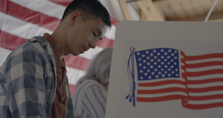 Low viewpoint, profile, looking up at Asian man voting at booth while considering candidates in United States election. Moves from medium shot to close up. Senior woman voter and US flag behind