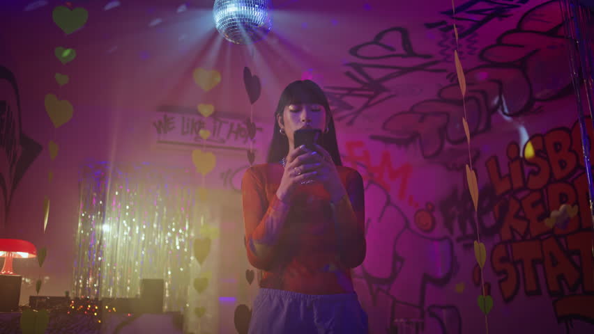 Clubbing girl looking smartphone engaged in vibrant club ambiance. Asian female clubber browsing mobile phone application at night party. Fashionable woman texting cellphone at neon light nightclub.