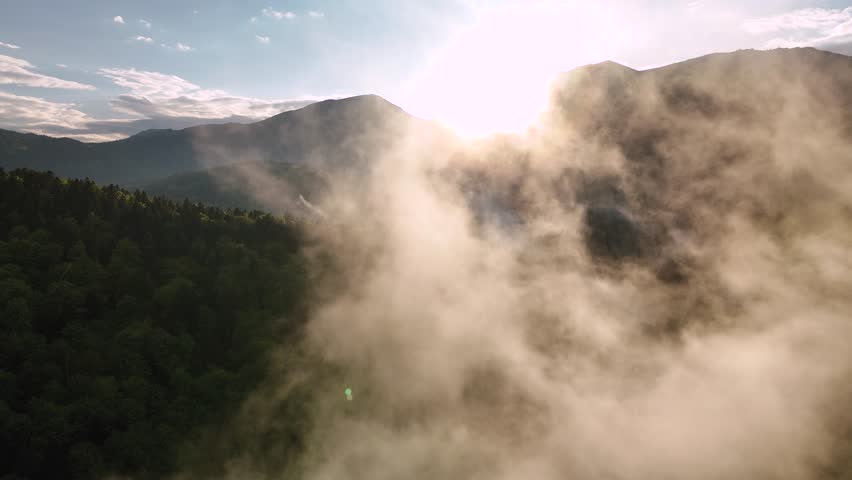 Flying trough fog at sunset unveiling mountains and forest landscape in late spring season. Aerial drone view of mist rising from woods in a rainy day