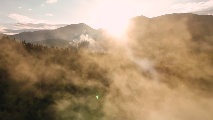 Flying trough fog at sunset unveiling mountains and forest landscape in late spring season. Aerial drone view of mist rising from woods in a rainy day.