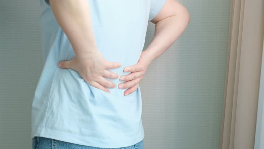 Young woman holding her lower back with her hands against wall background rear view back pain concept.