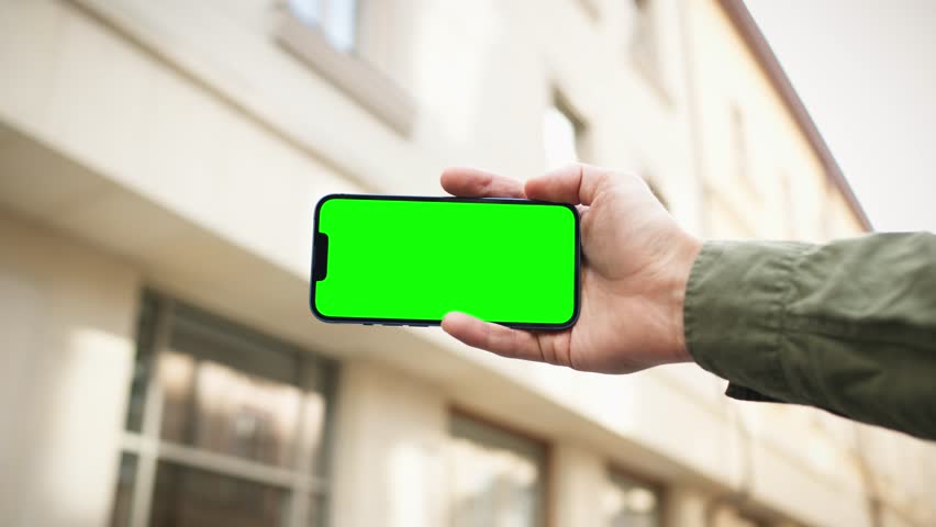 Camera focus on smartphone with green screen. Someone holding mobile device horizontally with one hand. Chroma key visible on display. While building in blurred background. Sunny weather.