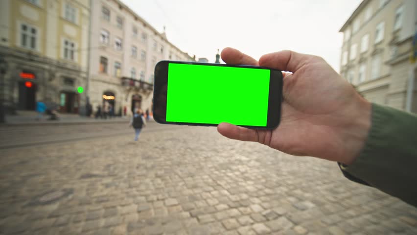 Close camera view of smartphone with chroma key. Someone holding mobile device horizontally with one hand. Standing in middle of city centre. Gadget with green screen on display. Concept of technology