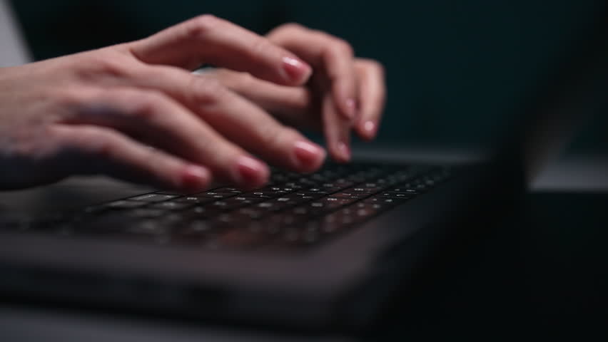 Closeup of hands of unrecognizable female hacker programmer working typing on keyboard laptop computer in dark room. Freelancer using laptop working from home office. Shooting in slow motion.