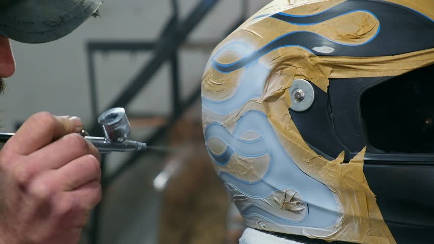 Handsome bearded, mustachioed biker side view intently painting wavy pattern on motorcycle helmet with white paint spray.Paint technique using masking unpainted surface with piece of paper and tape.
