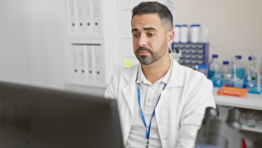 Furious young hispanic man in scientist uniform, shouting loud at computer in lab, hand over mouth amplifying his angry scream