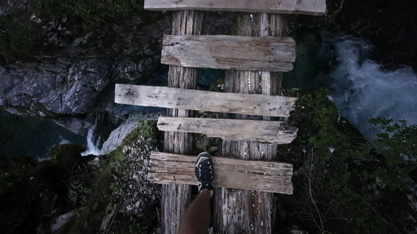 POV of a male hiker walking across an old wooden bridge with a waterfall and rocks beneath at Blue Eye of Theth in Albania