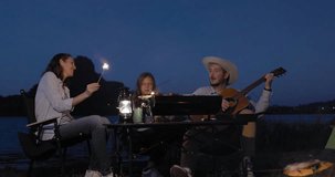 Happy family backpacker eating BBQ and drink and playing guitar together with happiness in Summer while having camping near lake at twilight time, 4k video