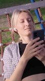 Caucasian young blonde woman using smartphone, listening to music in headphones, in hammock in garden, relaxing and summer concept using internet or social media and browsing mobile app.