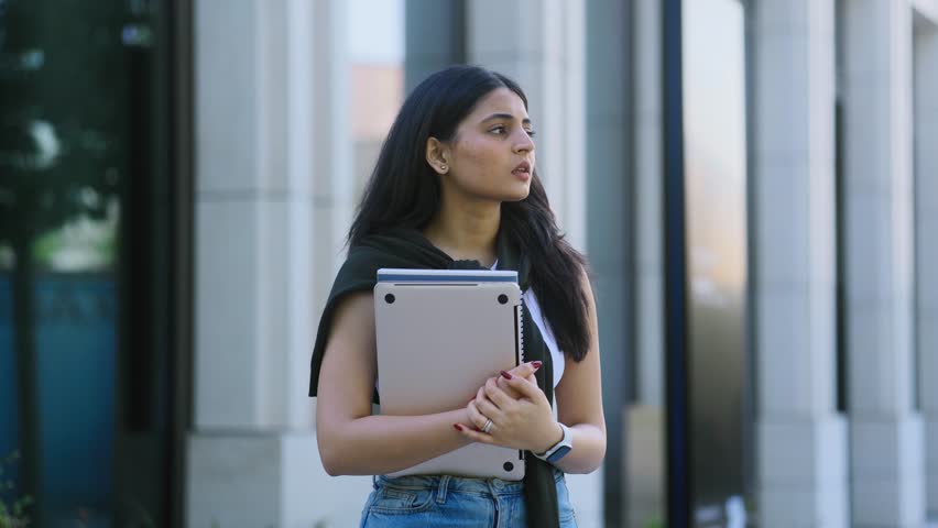 Sad young indian woman with notebooks waiting to meet with someone at city street Upset female looking at watch and looking around outdoors Lack of punctuality concept