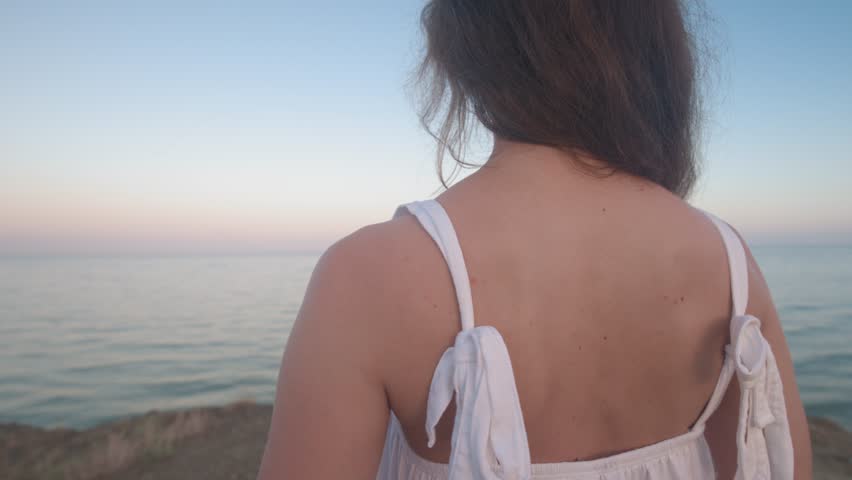 The back of a beautiful girl in a white dress with bare shoulders on a tropical beach. Long hair flutters in the wind against the background of the sea, a woman looks into the distance