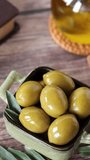 Pouring extra virgin olive oil on top of green olives with holy bible book, branch and leaves on rustic wooden table. Close-up. Vertical video.