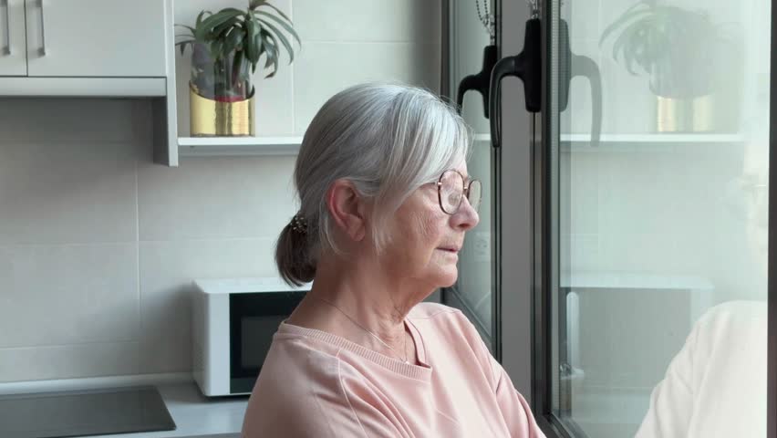 A sad elderly woman looks out the window expressing melancholy while her husband consoles her, an elderly married couple hugs and supports each other in a difficult time