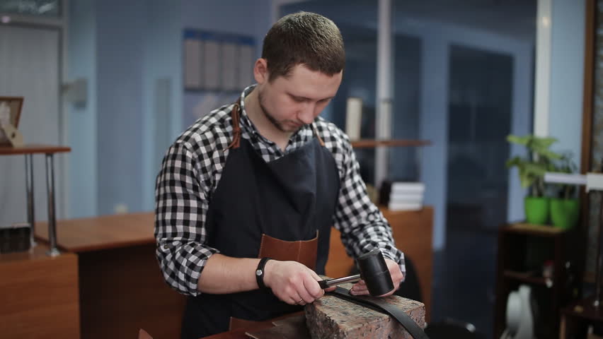 Working process of the leather belt in the leather workshop. Man holding crafting tool and working. He is placing billet to sturdy stand, fixing round knife on a mark and hitting with a small hammer Royalty-Free Stock Footage #35030956