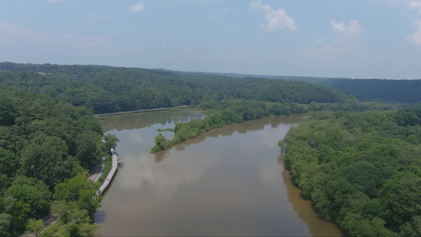 stunning aerial footage of the still green waters of the Chattahoochee river surrounded by lush green trees, grass and plants with blue sky and clouds at Roswell Riverwalk Boardwalk in Roswell Georgia