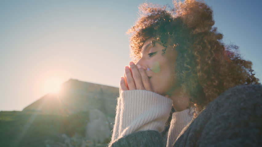Curly brunette enjoying sunset at wild nature portrait. Happy african american woman warming hands looking camera in warm sunlight