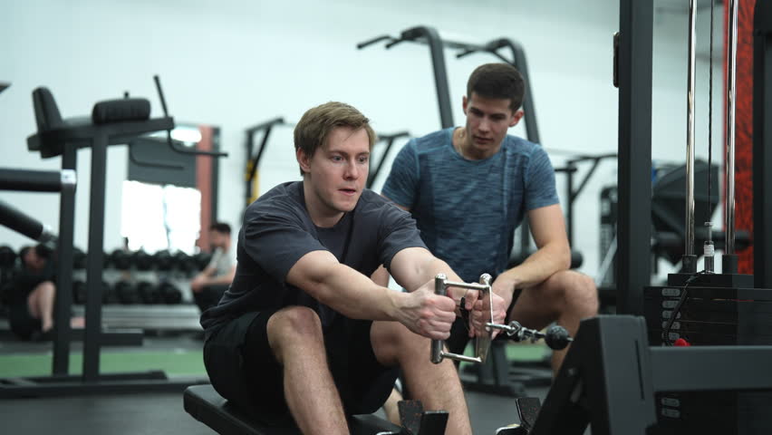 Beginner sportsman hard time practicing on rowing machine during sports training with personal coach in health club. Male athlete doing exercise with strap on rowing machine during fitness training