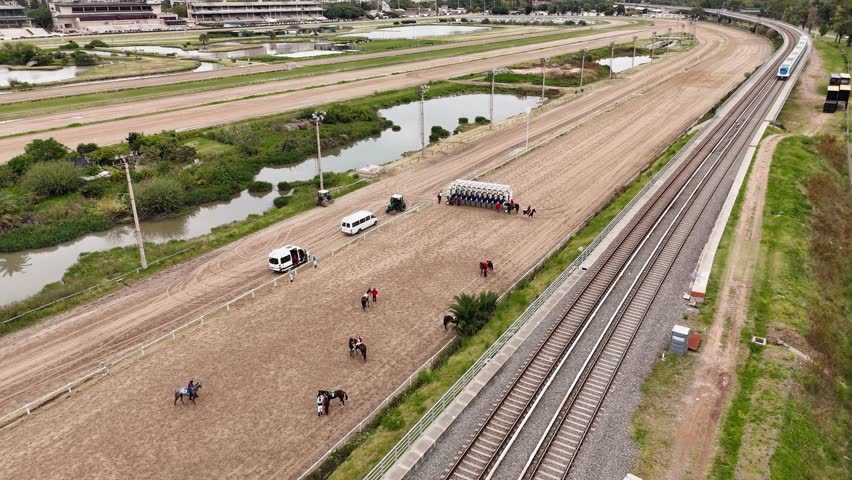Jockeys prepare their horses to start at the Palermo Racecourse (Buenos Aires, Argentina) and a train passes by. Static aerial camera