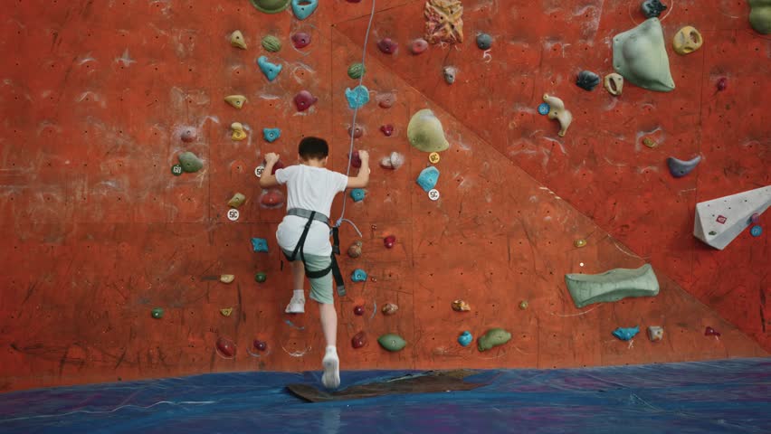 The main plan, the camera zooms in on a 10-year-old boy who starts climbing the wall of a climbing wall indoors. A small purposeful brave boy climbs to a great height on a climbing wall. 4k footage Royalty-Free Stock Footage #3503403675