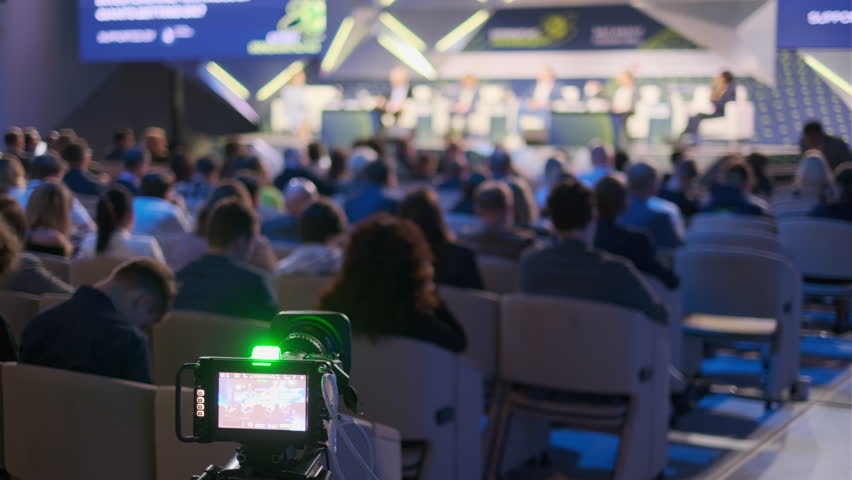 An image capturing a business conference from the perspective of a professional video camera, focusing on blurred audience and speakers.
