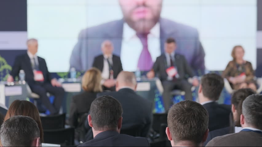 Focused audience at an international conference listening to speakers discussing global issues, with a blurred forefront and sharp background.
