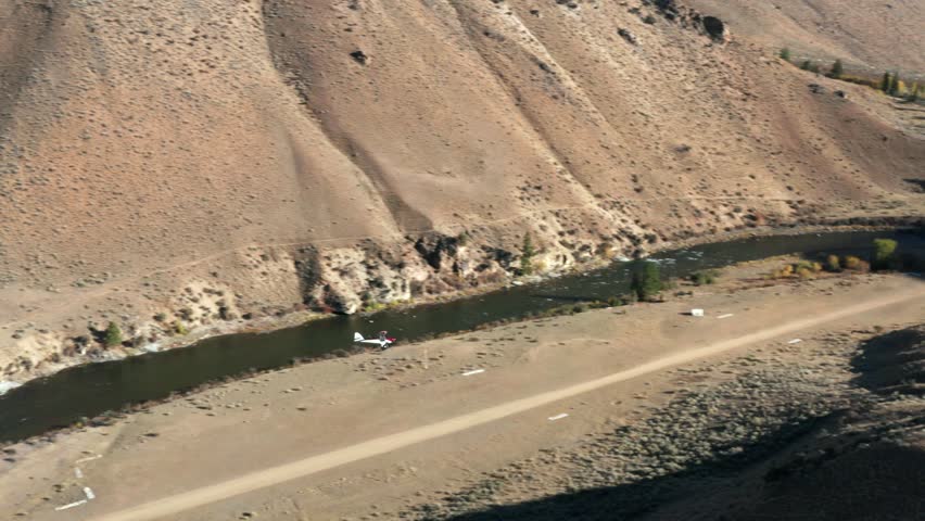 Drone footage from the side of a backcountry single-engine plane landing on a remote landing strip surrounded by mouintains and a river in the Frank Church River of No Return Wilderness in Idaho