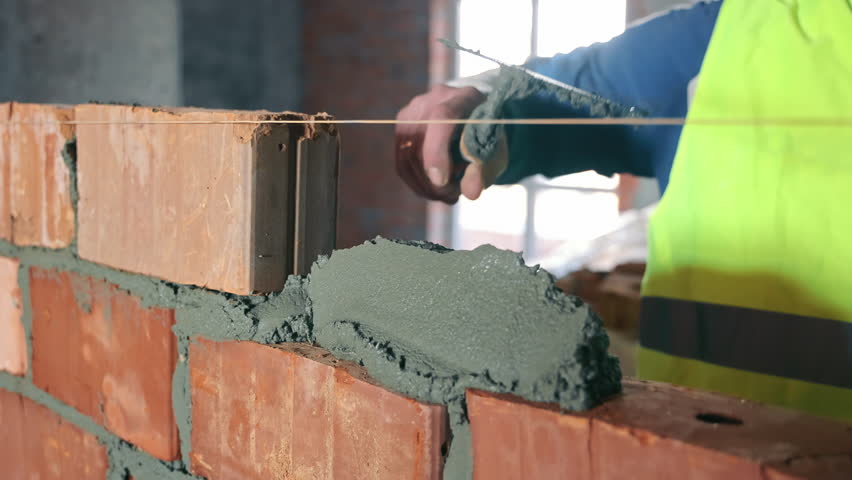 Close-up of Mortar Application on Brick Wall, Close-up of a construction worker's hands applying mortar on a brick wall, demonstrating precision in bricklaying techniques.