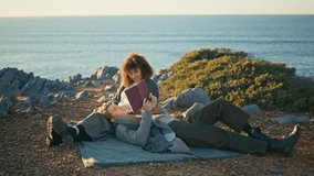 Relaxed young couple lying on picnic blanket at marine nature. Attractive blonde man reading book loud to happy woman in autumn ocean beach. Carefree family chilling on hill. Weekend leisure concept  - Powered by Shutterstock - Get 15% off with code: PIKWIZARD15