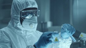 African American scientist in protective coverall, face mask, gloves and glasses pouring liquid from test tube into flask with biological hazard sticker during laboratory test - Powered by Shutterstock - Get 15% off with code: PIKWIZARD15
