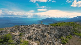 A couple woman and man stand on mountain peak overlooking the Beagle Channel at windy day in Ushuaia, Tierra del Fuego, Argentina. Aerial drone video.