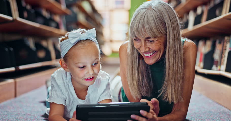 Mature woman, girl and happy with tablet in library on floor for educational games or ebook for child development. Grandmother, kid and smile for teaching or learning with support, bonding and care