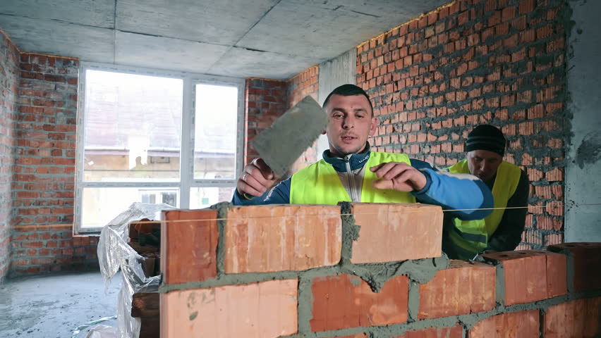 Construction Worker Building a Brick Wall, A focused construction worker in a reflective vest meticulously lays bricks on a partially built wall inside a building under renovation.