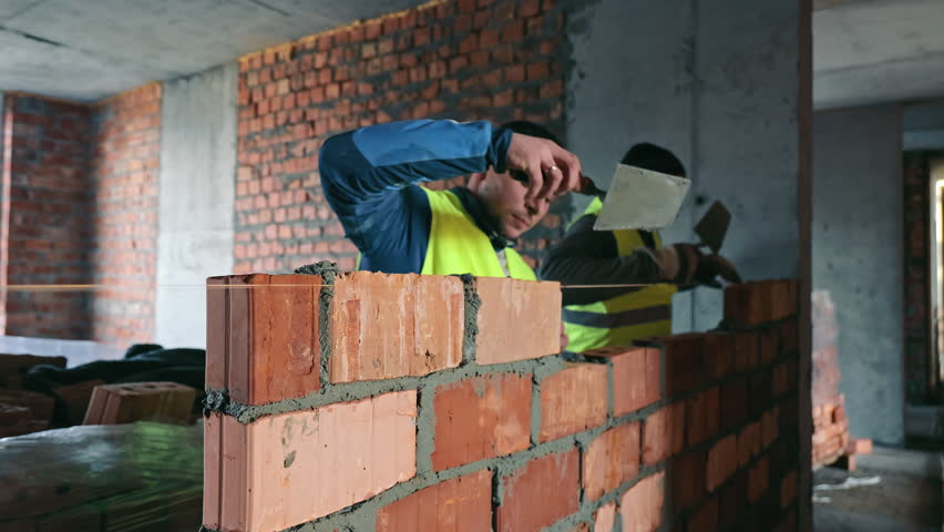 Construction Worker Building a Brick Wall, A focused construction worker in a reflective vest meticulously lays bricks on a partially built wall inside a building under renovation.