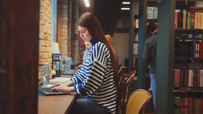 Woman focuses on laptop while taking notes in cozy cafe with brick walls and warm lighting. Productive work environment with relaxed atmosphere, blending concentration and comfort - Powered by Shutterstock - Get 15% off with code: PIKWIZARD15