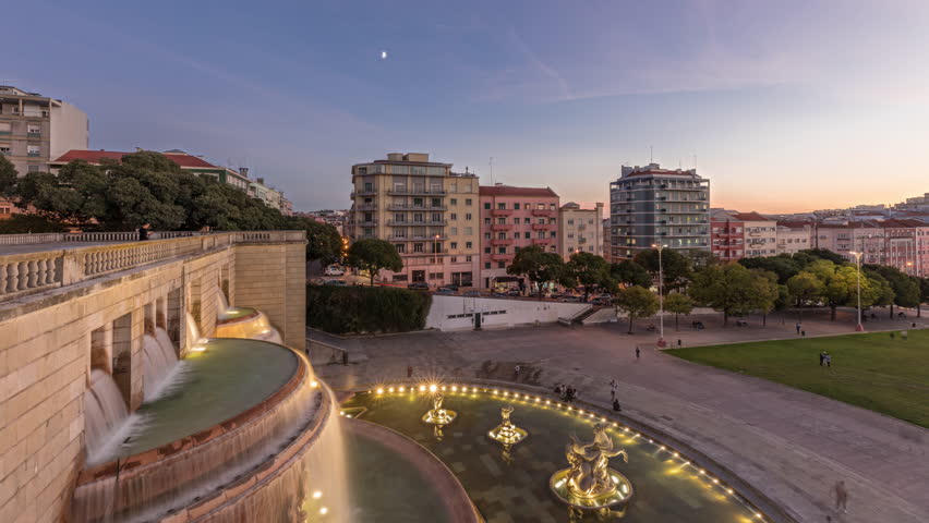 Panorama showing lawn at Alameda Dom Afonso Henriques with colorful buildings and illuminated Luminous Fountain aerial day to night transition timelapse from above after sunset in Lisbon, Portugal