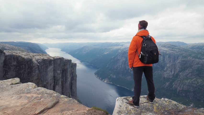 Side view from behind of tourist in warm clothes stands on top of rock, enjoying the beautiful natural landscape in Norway. Full Shot. Young sporty man is inspired by magnificent view of the landmark