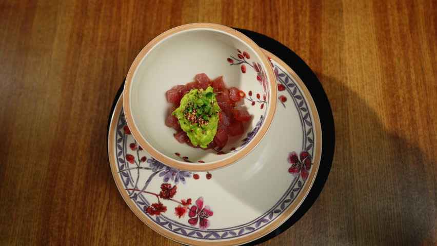 Fancy presentation of raw tuna tartare combined with creamy avocado sauce enhancing taste of seafood. Delicious appetizer made of uncooked fish served in ceramic bowl on plate with floral ornaments.