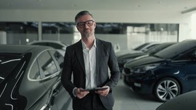 Portrait of a confident middle-aged man manager in a business suit with glasses and with a tablet in his hands who walks along modern black cars in a car showroom and takes inventory - Powered by Shutterstock - Get 15% off with code: PIKWIZARD15