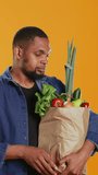 Vertical Video Vegan adult presenting a ripe carrot from his groceries paper bag, recommending local farming with ethically sourced eco friendly produce. Pleased guy showing a fresh vegetable. Camera