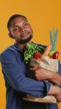 Vertical Video Male model hugging his paper bag full of organic homegrown food, enjoying heathy eating from local bio supermarket. Young vegan person recommending ethically sourced produce. Camera B.
