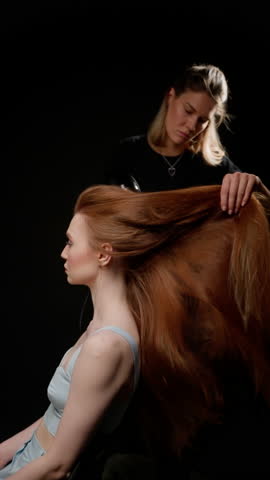 Profile of a young woman with long red hair being blow-dried by a hairstylist against a black background