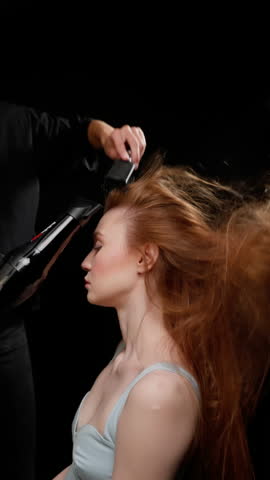 Side view of a young woman with long red hair being blow-dried by a hairstylist in a dark studio setting