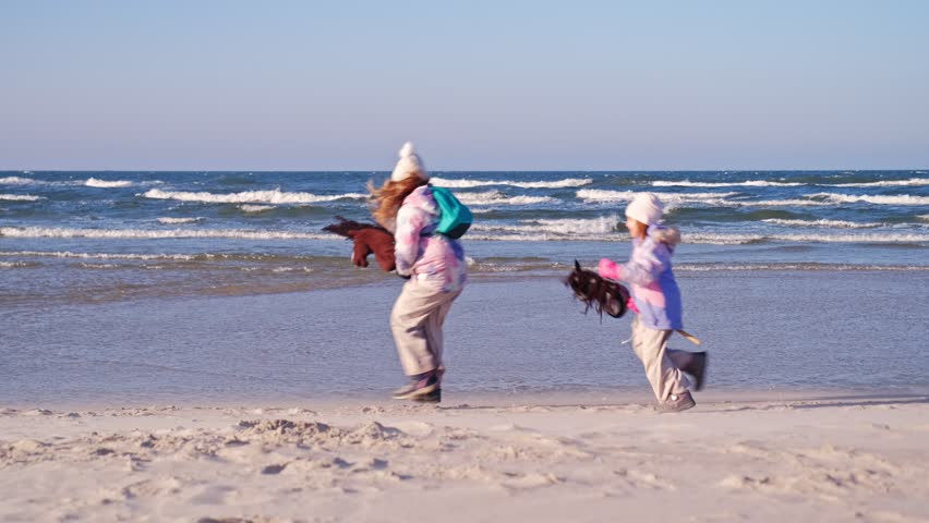 Two Happy Caucasian Girls Playing with Hobby Horse on Stick Toy on Seashore Beach on Windy Day