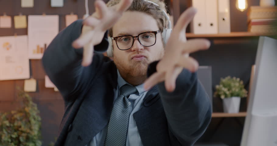 Loop of joyful businessman dancing listening to music with headphones moving hands and looking at camera in office. Businessperson and youth culture concept.