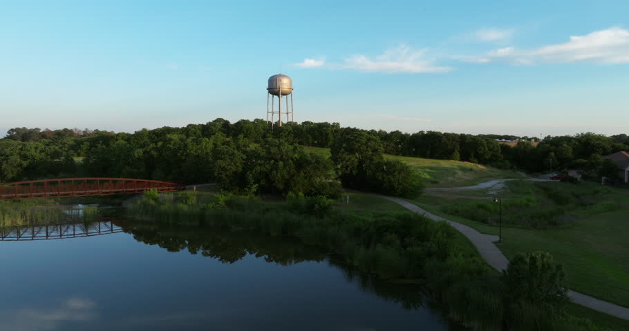 Cinematic dusk aerial of a picturesque local park in a new suburban development with a reflection pond and a pedestrian bridge with people with green trees, grass, and a water tower in the background.
