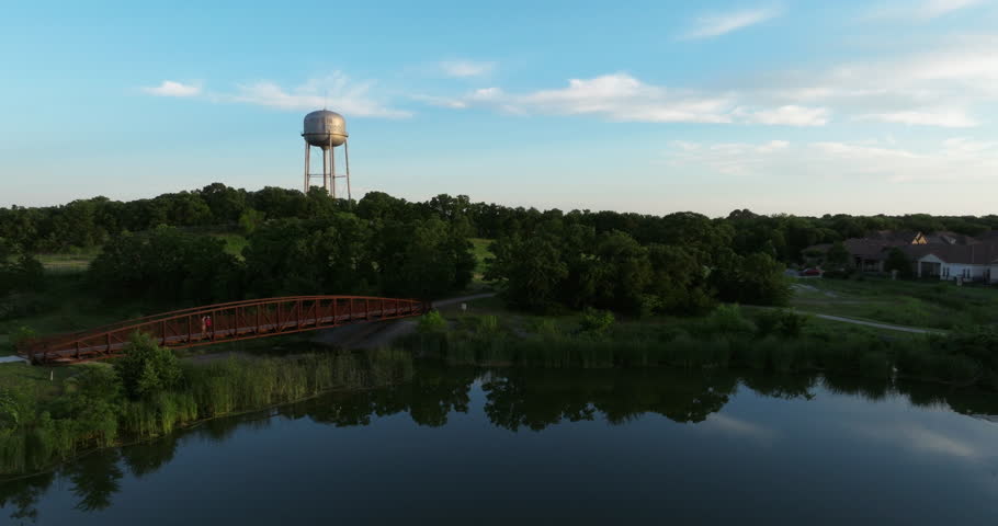 Real Estate setting shot of a local park with a beautiful reflective pond near a modern metal pedestrian bridge, green trees and grass, blue sky, and a picturesque water tower on a hill at dusk.