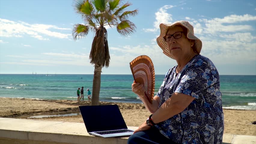 Slow motion, elderly woman fanning herself sitting next to a laptop, near the beach, on a hot summer day.