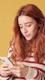 Vertical video, Surprised red-haired woman dressed in sweater, looking at camera while holding smartphone on yellow background