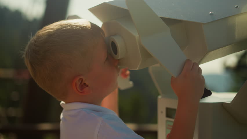 Inquisitive boy explores world via massive binoculars. Little child in white polo enjoys wonderful view of undulating mountains standing on observation deck